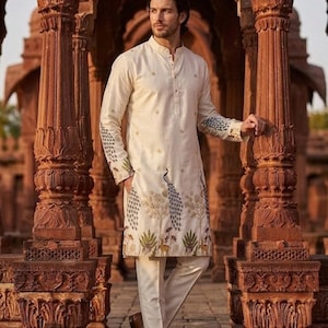May include: An off-white long-sleeved kurta tunic featuring a mandarin collar and button placket, adorned with embroidered peacock and floral motifs along the hem and sleeves, paired with matching off-white trousers and brown leather loafers.