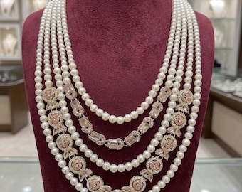 Könnte beinhalten: Eine mehrsträngige Perlenkette mit roségoldenen und kristallenen Akzenten. Die Halskette wird auf einer weinroten Büste präsentiert. Das Design umfasst florale und geometrische Elemente, mit einem rechteckigen Anhänger unten.