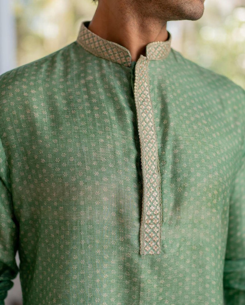 Puede incluir: Una camisa verde con estampado y un ribete bordado blanco y plateado alrededor del cuello y en la parte delantera.