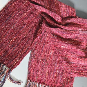 May include: A woven scarf in shades of red with a textured appearance. The scarf has a fringed edge with strands of various colors, including blue, orange, and purple. The fabric appears soft and drapes loosely.
