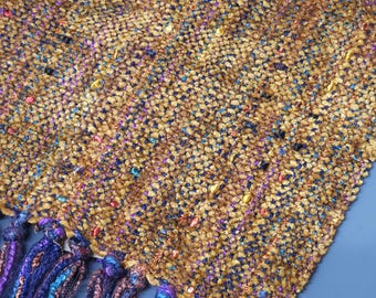 Könnte beinhalten: Ein handgewebter Schal mit einem strukturierten, gesprenkelten Muster in Gold-, Braun- und Blautönen. Der Schal hat einen Fransenbesatz aus dickem, gezwirntem Garn in Lila, Blau und Braun.