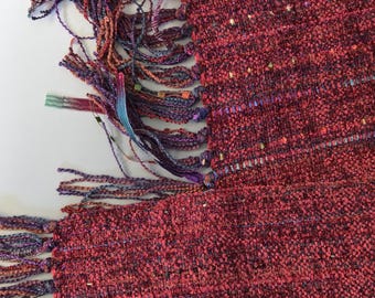 Könnte beinhalten: Ein gewebter Schal aus tiefrotem und burgunderfarbenem strukturiertem Stoff mit dezenten blauen und violetten Sprenkeln. Die Kanten sind mit langen, geknoteten Fransen in einer bunten Mischung aus violetten, blauen und roten Garnen versehen.