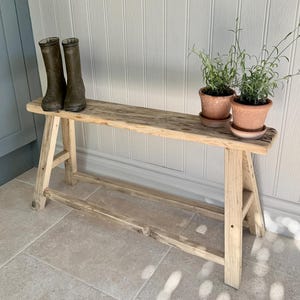 May include: A reclaimed wooden bench with a natural, aged finish. Two small terracotta pots with plants and a pair of green wellington boots are on the bench. The bench is supported by angled legs, and is approximately 100cm long.
