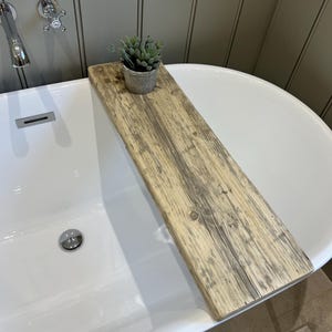 Puede incluir: Una bandeja de bañera de madera rústica con una pequeña planta suculenta en maceta. La bandeja, hecha de madera envejecida, abarca una bañera blanca. La planta está en una pequeña maceta de color plateado.