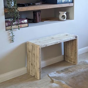 Puede incluir: Banco de madera rústico con un acabado claro y envejecido. El banco tiene un diseño simple y rectangular con un asiento plano y cuatro patas resistentes. El banco se coloca frente a un nicho de pared con libros y objetos decorativos.