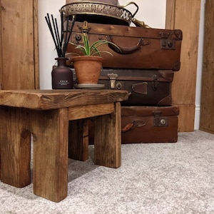 May include: A rustic wooden stool with a unique arched design, made from reclaimed wood. The stool is sitting on a grey carpet in front of a stack of vintage suitcases.