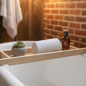 May include: A light wood bathtub tray holds a rolled white towel, a small succulent in a gray pot, and a brown bottle. The tray rests on the edge of a white bathtub, with a brick wall in the background.
