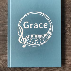 May include: A blue hardback journal with a white design of a treble clef, piano keys, and music notes. The word "Grace" is written in a circle.