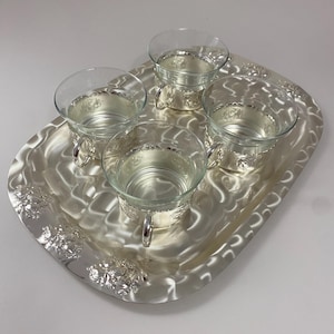 Può includere: Un vassoio d'argento con quattro tazze di vetro trasparente. Il vassoio presenta un motivo floreale decorativo sui bordi e una superficie strutturata. Le tazze hanno basi e manici argentati.