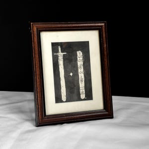 May include: Framed artwork featuring a detailed illustration of a sword and its scabbard. The artwork is in black and white, with intricate designs on both the sword and scabbard. The frame is a dark brown color, adding a classic touch to the piece.
