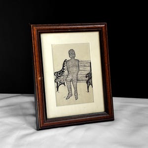 May include: Framed artwork featuring a pen and ink drawing of a figure sitting on a park bench. The figure is rendered in a textured style, with a dark brown wooden frame and a cream-colored mat.