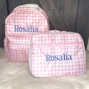 May include: A pink and white gingham backpack and matching lunch bag. Both items have the name "Rosalia" embroidered in blue. The backpack has a white handle and the lunch bag has a ruffle detail.