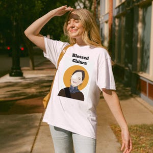 Puede incluir: Camiseta blanca con un gráfico de una mujer con un halo amarillo y el texto "Blessed Chiara" impreso en la parte delantera.