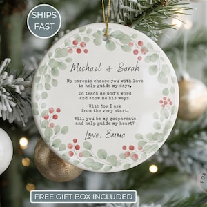 May include: A white ceramic Christmas ornament with a gold string, featuring a floral design with red berries and green leaves. The ornament has text that reads "Michael & Sarah" and a request to be godparents.