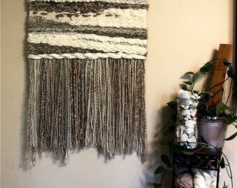 May include: A woven wall hanging with a brown and white color scheme. The hanging has a fringe of yarn at the bottom.