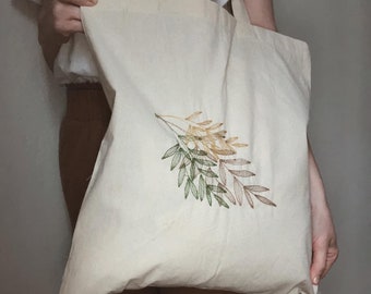 May include: A white canvas tote bag with a green, yellow, and brown embroidered leaf design.