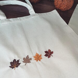 May include: A white canvas tote bag featuring four embroidered autumn leaves in shades of brown, orange, and yellow. The leaves are arranged in a row across the bottom of the bag.