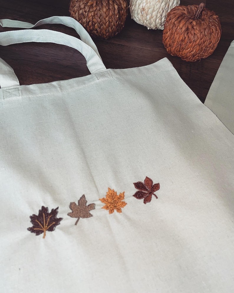 May include: A white canvas tote bag featuring four embroidered autumn leaves in shades of brown, orange, and yellow. The leaves are arranged in a row across the bottom of the bag.