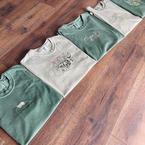 May include: Five folded green and beige t-shirts with different nature-themed designs. The designs include a sheep, a sun, and floral patterns. The shirts are laid out on a wooden surface.