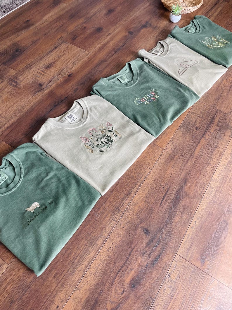 May include: Five folded green and beige t-shirts with different nature-themed designs. The designs include a sheep, a sun, and floral patterns. The shirts are laid out on a wooden surface.