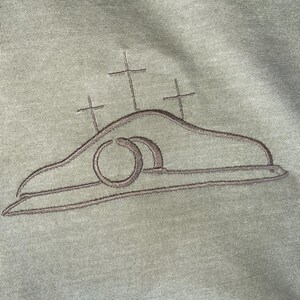 May include: A green t-shirt with an embroidered design of a tomb with a stone rolled away and three crosses above it.