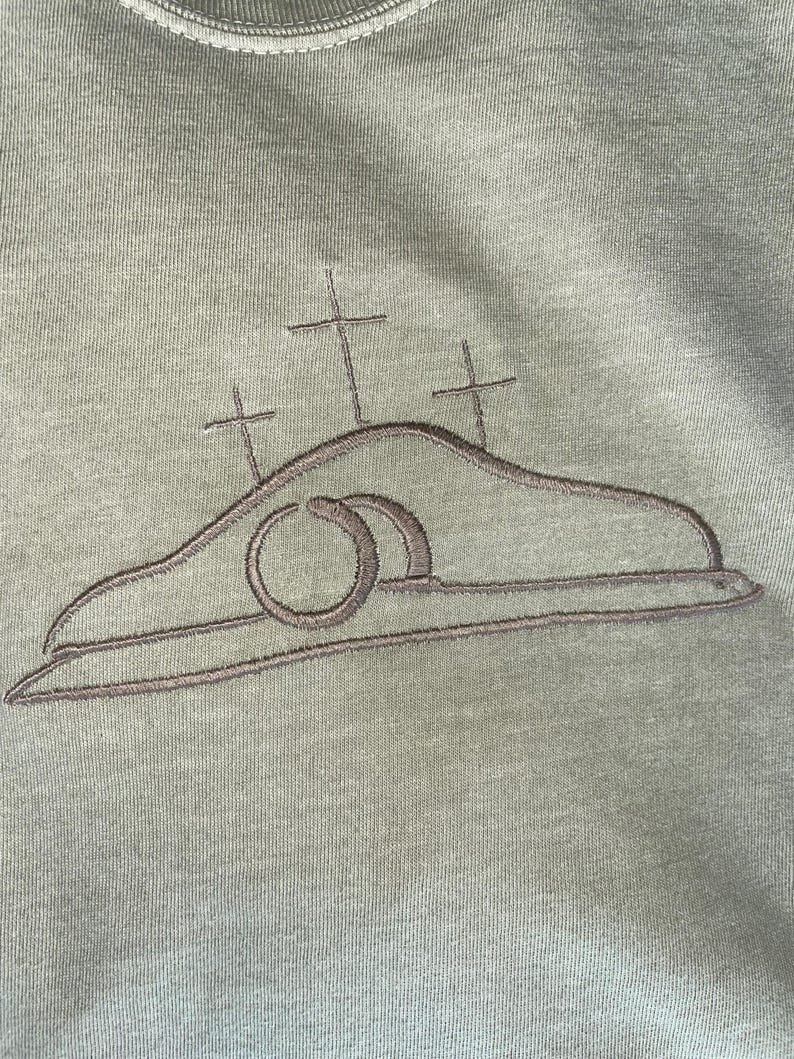 May include: A green t-shirt with an embroidered design of a tomb with a stone rolled away and three crosses above it.