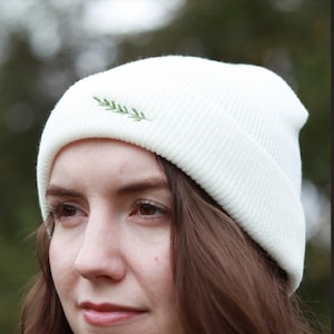 May include: A white beanie with a small green embroidered leaf design. The beanie is made of a soft, knitted fabric.