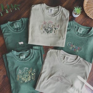 May include: Five different t-shirts with embroidered designs. The shirts are in shades of green and beige. The designs include flowers, a lamb, a cross, and the words "Made for Sunny Days".