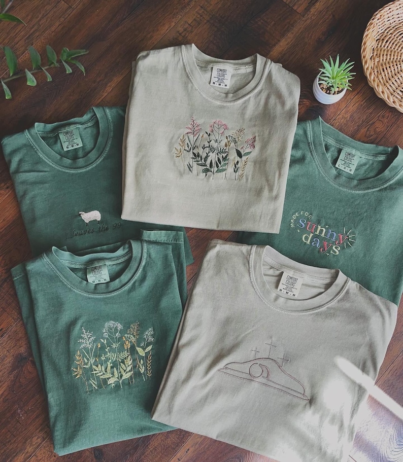 May include: Five different t-shirts with embroidered designs. The shirts are in shades of green and beige. The designs include flowers, a lamb, a cross, and the words "Made for Sunny Days".