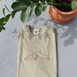 May include: A light green t-shirt with an embroidered image of a tomb with a cross on top. The t-shirt has a Comfort Colors tag on the bottom left side of the front.