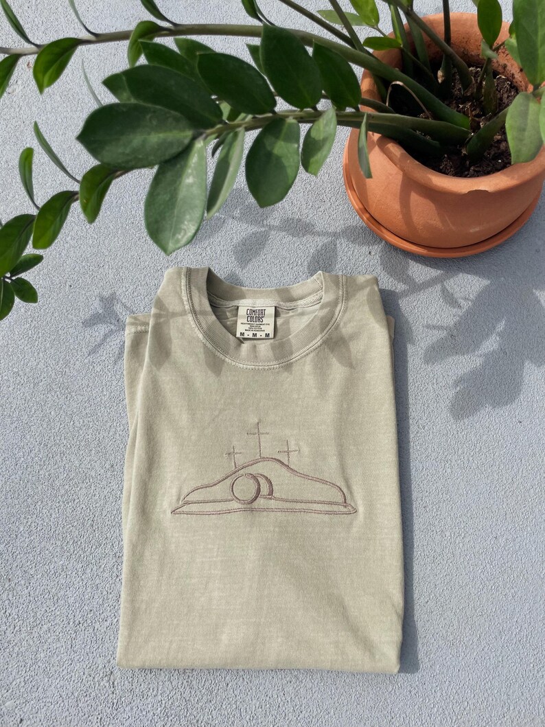 May include: A light green t-shirt with an embroidered image of a tomb with a cross on top. The t-shirt has a Comfort Colors tag on the bottom left side of the front.