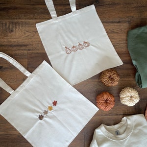 May include: Two white tote bags with embroidered designs. One bag features a line of five pumpkins, the other features a line of five autumn leaves in various shades of brown, orange, and yellow. A white sweatshirt with an embroidered line of five pumpkins is also pictured.