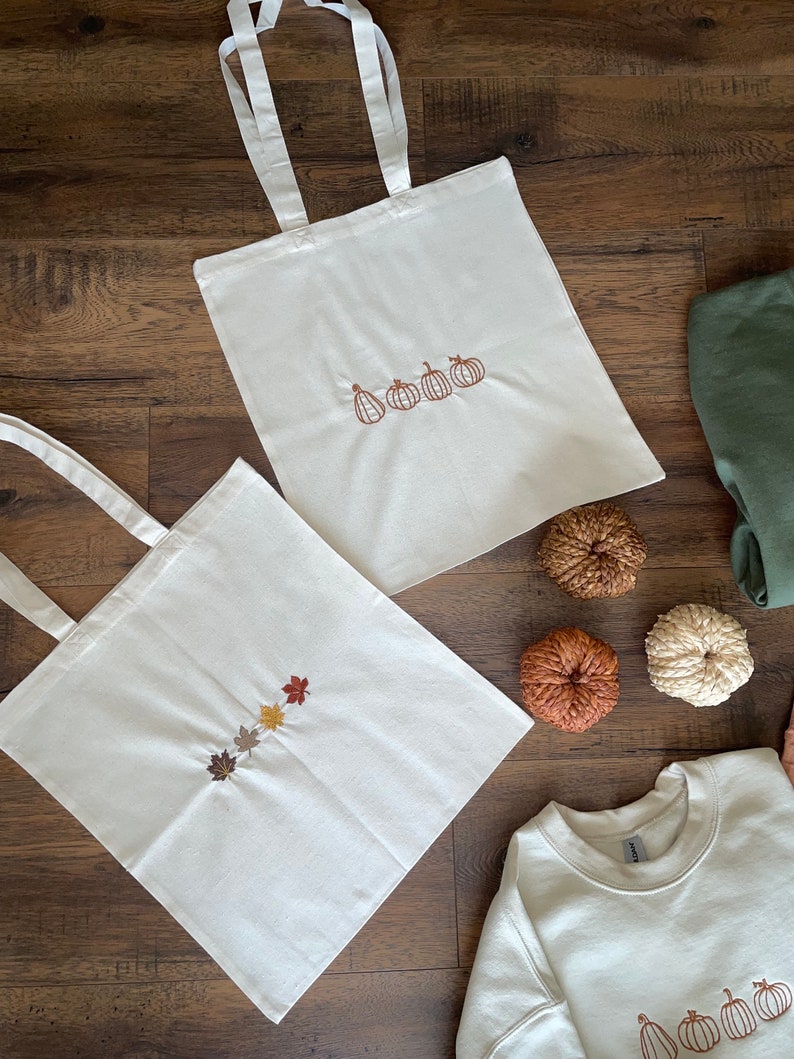 May include: Two white tote bags with embroidered designs. One bag features a line of five pumpkins, the other features a line of five autumn leaves in various shades of brown, orange, and yellow. A white sweatshirt with an embroidered line of five pumpkins is also pictured.
