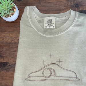 May include: A light green sweatshirt with an embroidered design of three crosses and a stone tomb. The text "COMFORT COLORS" is on a tag attached to the sweatshirt.