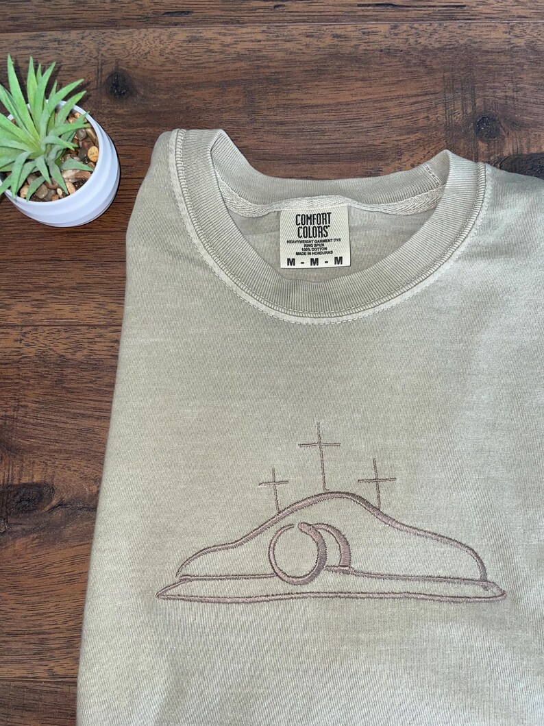 May include: A light green sweatshirt with an embroidered design of three crosses and a stone tomb. The text "COMFORT COLORS" is on a tag attached to the sweatshirt.