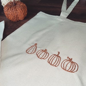 May include: A white canvas tote bag with brown embroidered pumpkins. The pumpkins are arranged in a row across the bottom of the bag.