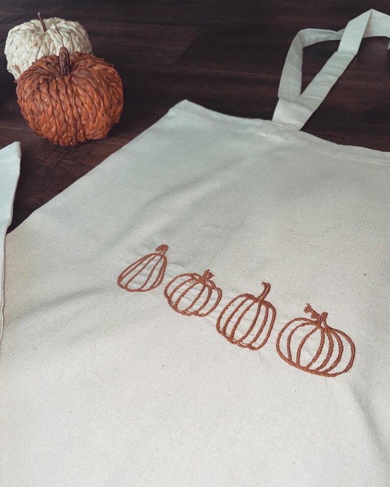 May include: A white canvas tote bag with brown embroidered pumpkins. The pumpkins are arranged in a row across the bottom of the bag.