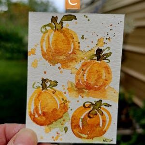 May include: A watercolor painting featuring four orange pumpkins with green stems and leaves. The pumpkins are painted with loose brushstrokes and surrounded by orange and green paint splatters. The letter "C" is in the upper left corner.