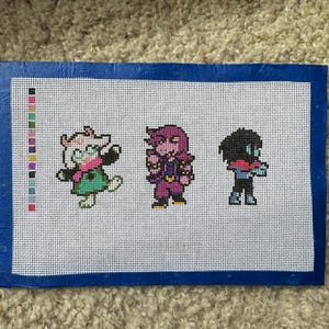 May include: A rectangular piece of white cross-stitch fabric featuring three pixelated character figures in a row. To the left is a vertical color palette guide. The fabric is bordered by a dark blue tape frame on all four sides of the canvas.