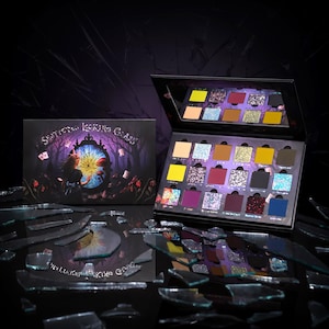 May include: An open eyeshadow palette with a variety of colours, including yellow, pink, purple, and blue. The palette is next to a box with the text "Shattered Looking Glass" and an illustration of a girl looking into a mirror. The background is dark with broken glass.