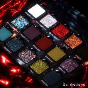 Pode incluir: Uma paleta de sombras preta com 16 sombras em forma de diamante. As tonalidades incluem prata brilhante, vermelho e dourado, juntamente com verde-azulado fosco, bordô e amarelo. A paleta está sobre um fundo vermelho texturizado. O texto na paleta inclui "Twisted Rabbit" e "Rabbit Hole."