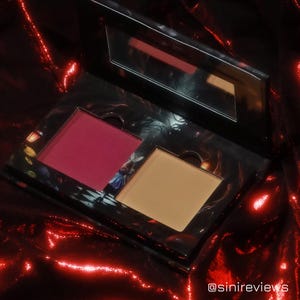 Può includere: Una palette di trucco compatta con due tonalità di blush. La palette ha uno sfondo nero con un'illustrazione fantasiosa di una scena di bosco. Le tonalità di blush sono un rosso intenso e una pesca chiara.