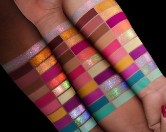 May include: Three forearms display multiple rectangular swatches of eyeshadow in a vibrant spectrum of colors. The palette includes matte and shimmer finishes in shades of deep burgundy, bright orange, hot pink, yellow, teal, and iridescent glitter.