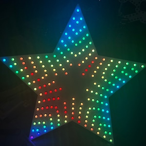 Puede incluir: Una estrella blanca de cinco puntas con luces LED de arcoíris. Las luces están dispuestas en un patrón circular alrededor de la estrella.