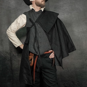 May include: A historical-style outfit featuring a black wide-brimmed hat, a cream-colored long-sleeve shirt with a ruffled collar, a dark gray button-up vest, a black capelet, black breeches, a brown leather belt, and beige stockings.