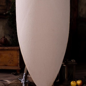 Op de afbeelding: Een groot, schildvormig verdedigingsmiddel met een effen, lichtbeige, getextureerd oppervlak. Het schild heeft een gebogen bovenrand die taps toeloopt naar een punt aan de onderkant. Het oppervlak lijkt bedekt met een textielachtig materiaal.