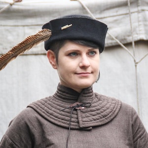 Puede incluir: Un sombrero de fieltro negro con un alfiler dorado y una pluma marrón. El sombrero se usa con un cuello acolchado marrón y una camisa marrón de manga larga. La persona de la imagen tiene el pelo corto y oscuro y mira hacia la derecha.