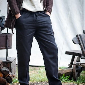 Könnte beinhalten: Eine Person trägt eine braune Steppjacke über einem cremefarbenen Hemd und einer schwarzen Hose. Die Hose hat einen Kordelzug in der Taille. Die Person trägt braune Lederschuhe. Die Umgebung scheint im Freien zu sein.