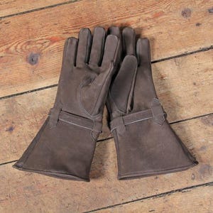 May include: A pair of brown leather gloves with long cuffs and a strap detail. The gloves are made of a soft, suede-like material and are laid out on a wooden surface. The gloves appear to be new and unworn.