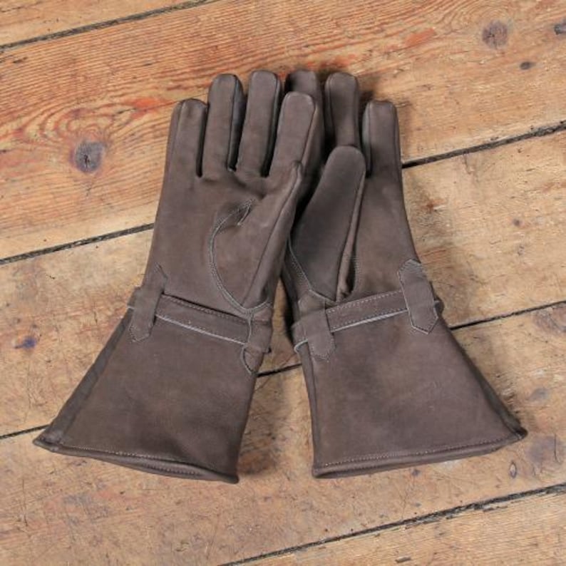 May include: A pair of brown leather gloves with long cuffs and a strap detail. The gloves are made of a soft, suede-like material and are laid out on a wooden surface. The gloves appear to be new and unworn.